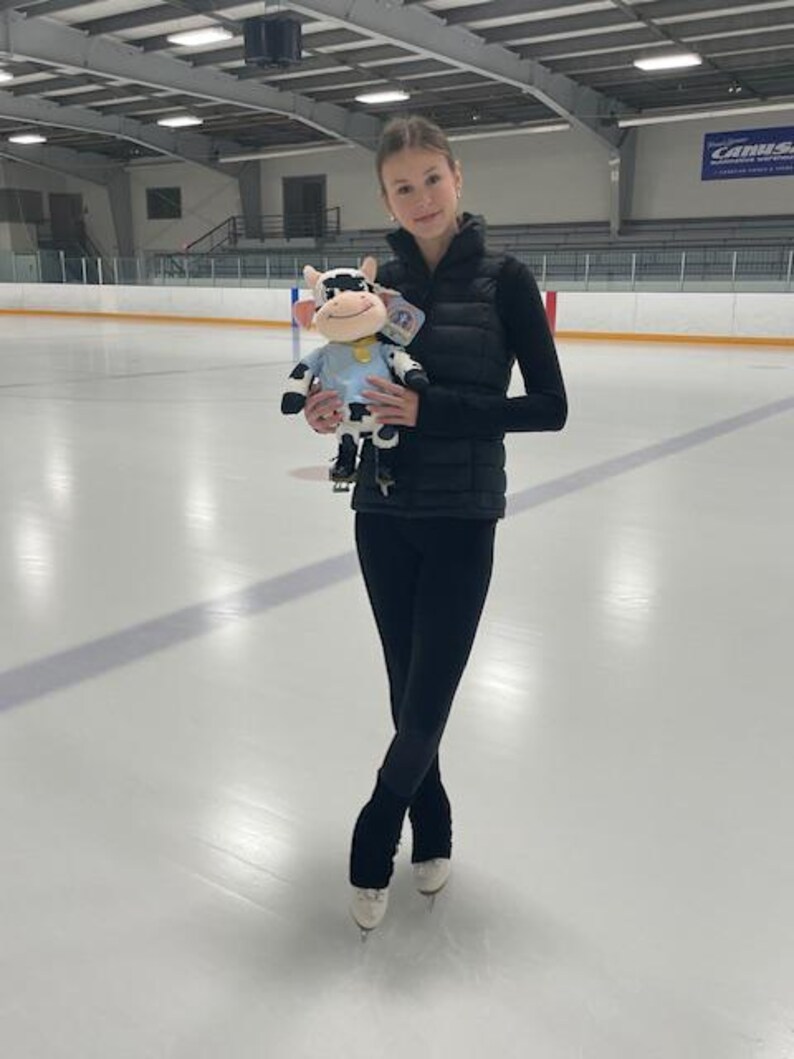 May include: A young person in black athletic wear and ice skates stands on an ice rink, holding a plush cow toy. The cow is white and black with a blue shirt and a yellow collar. The background shows an indoor ice rink.