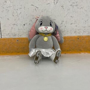 May include: A plush gray bunny wearing a gold medal, a white skirt, and ice skates. The bunny has pink inner ears, a pink nose, and is sitting on an ice rink. The bunny is a cute and cuddly toy.