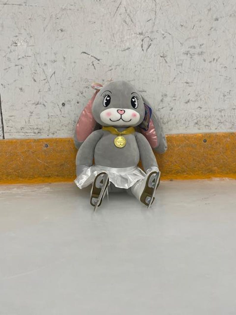 May include: A plush gray bunny wearing a gold medal, a white skirt, and ice skates. The bunny has pink inner ears, a pink nose, and is sitting on an ice rink. The bunny is a cute and cuddly toy.