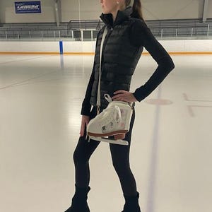 May include: A woman in black leggings and a black puffer vest stands on an ice rink, holding a pair of white ice skates with brown soles. The skates are in a white bag with a shoulder strap. She is wearing black skate guards.