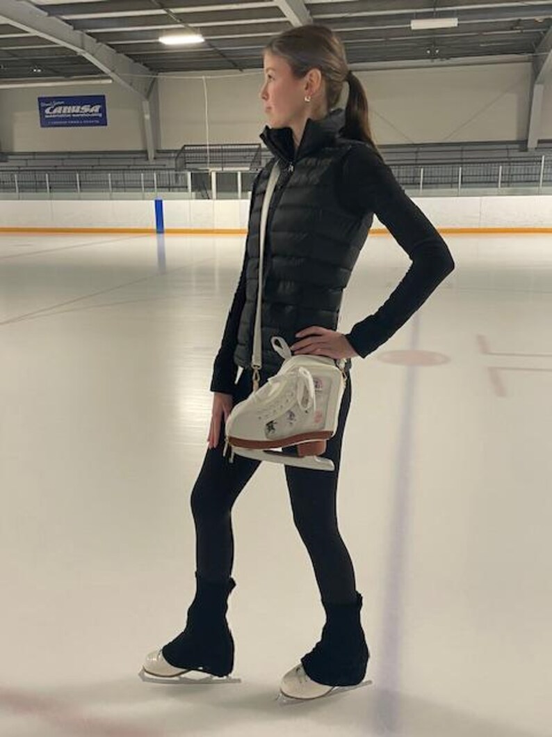 May include: A woman in black leggings and a black puffer vest stands on an ice rink, holding a pair of white ice skates with brown soles. The skates are in a white bag with a shoulder strap. She is wearing black skate guards.