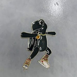 May include: A black frog-shaped pin with silver trim, wearing ice skates and a gold pendant. The frog has a shiny, glossy finish and is set against a gray background. The pin is a whimsical accessory.