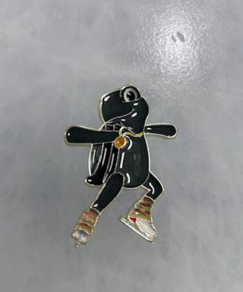 May include: A black frog-shaped pin with silver trim, wearing ice skates and a gold pendant. The frog has a shiny, glossy finish and is set against a gray background. The pin is a whimsical accessory.