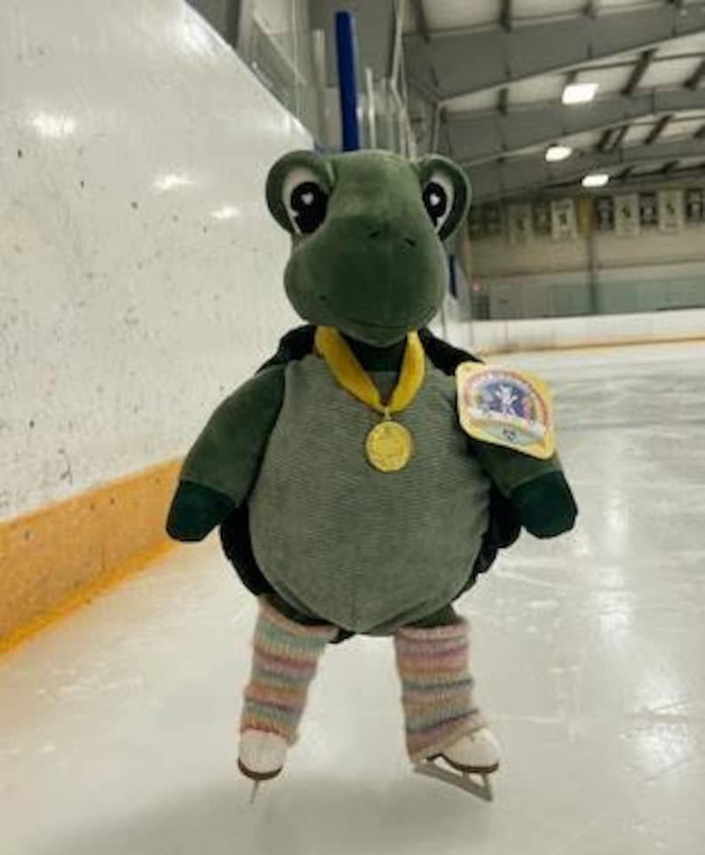 May include: A plush green turtle wearing a gold medal and striped leg warmers, posed on ice skates. The turtle has a black shell and a tag attached to its side. The background shows an ice rink.