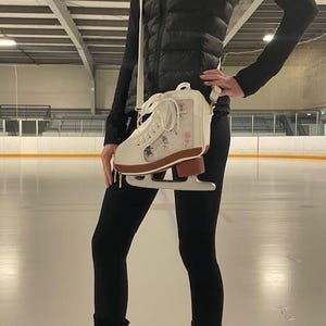 May include: A white ice skate-shaped crossbody bag with a brown blade, featuring floral designs and a clear panel. The bag is worn with a black puffer vest and leggings, set against an ice rink background.
