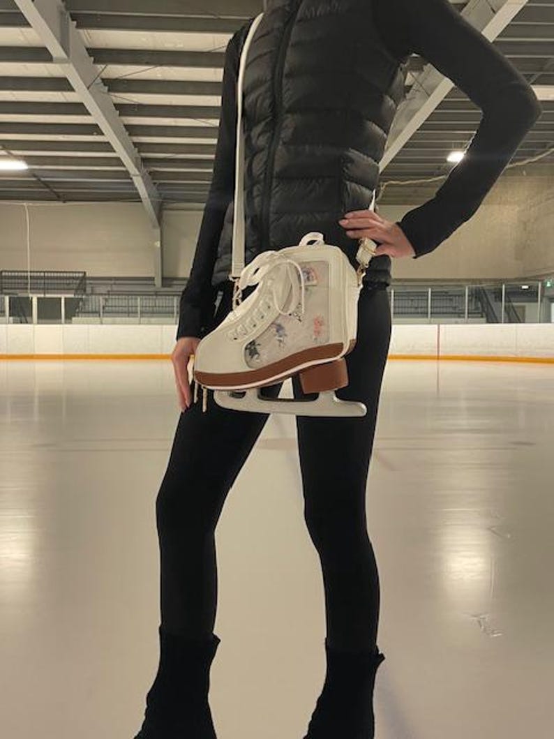 May include: A white ice skate-shaped crossbody bag with a brown blade, featuring floral designs and a clear panel. The bag is worn with a black puffer vest and leggings, set against an ice rink background.