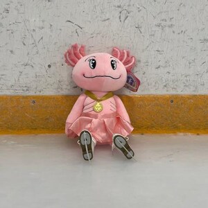 May include: A plush pink axolotl toy wearing a pink dress, gold medal, and ice skates. The axolotl has pink frills and a smiling face. The toy is sitting on an ice rink.