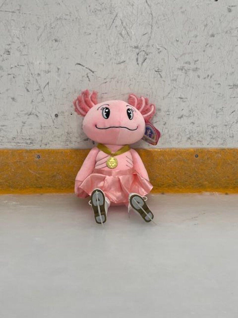 May include: A plush pink axolotl toy wearing a pink dress, gold medal, and ice skates. The axolotl has pink frills and a smiling face. The toy is sitting on an ice rink.