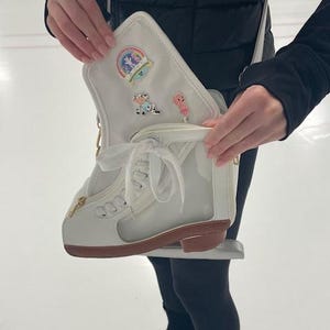 May include: A white ice skate-shaped purse with a brown sole and gold zipper. The purse has a clear side panel and is decorated with pins, including a rainbow and cartoon characters. The purse is held open, revealing the interior.