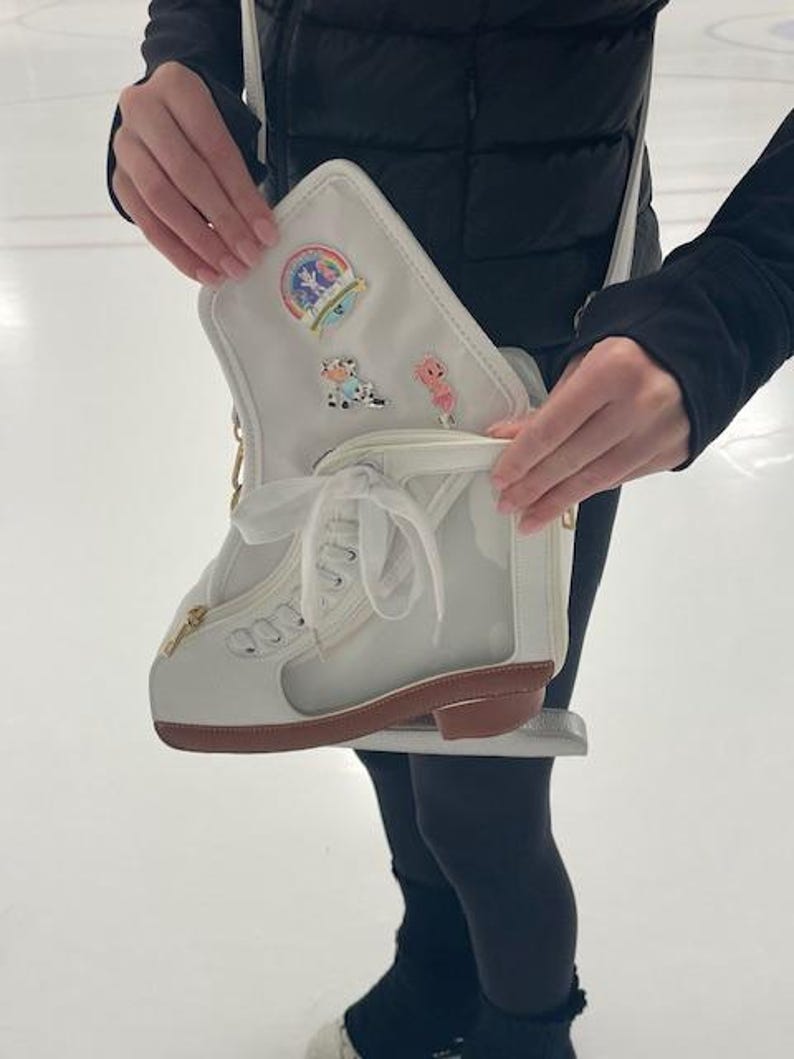 May include: A white ice skate-shaped purse with a brown sole and gold zipper. The purse has a clear side panel and is decorated with pins, including a rainbow and cartoon characters. The purse is held open, revealing the interior.