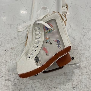 May include: White ice skate-shaped crossbody bag with a clear window displaying colorful pins. The bag has brown trim, a silver blade, and a white strap with gold-colored hardware. The laces are white.