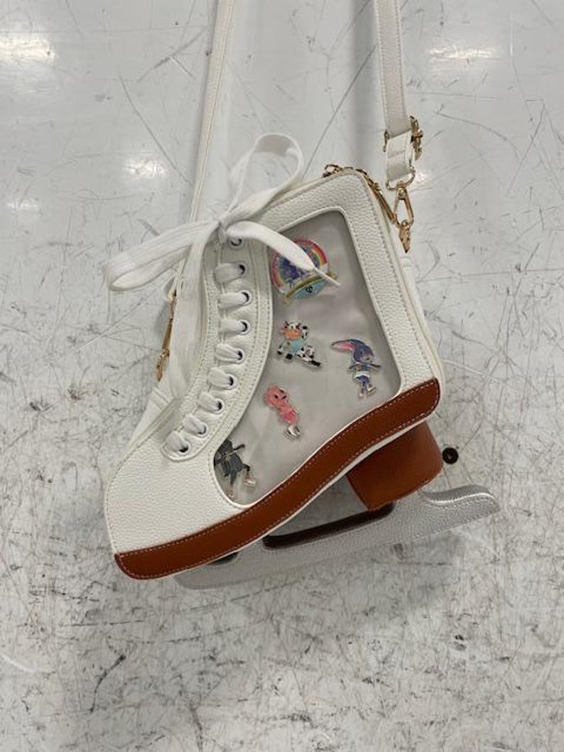 May include: White ice skate-shaped crossbody bag with a clear window displaying colorful pins. The bag has brown trim, a silver blade, and a white strap with gold-colored hardware. The laces are white.