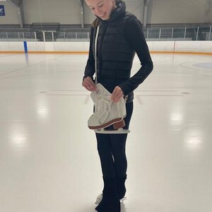 May include: A person stands on an ice rink, holding a pair of white ice skates with brown soles. The person is wearing black leggings, a black vest, and a black long-sleeved shirt. The background shows the ice rink and the ceiling.