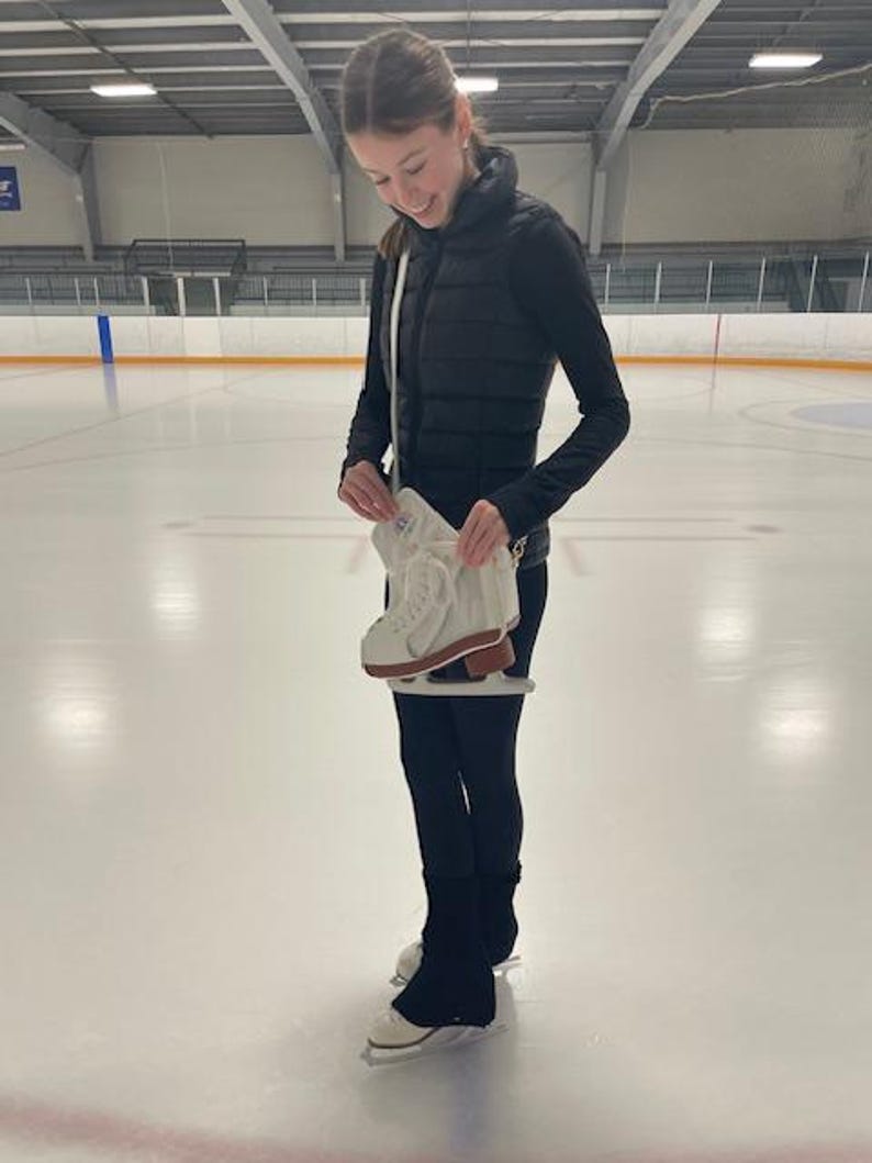 May include: A person stands on an ice rink, holding a pair of white ice skates with brown soles. The person is wearing black leggings, a black vest, and a black long-sleeved shirt. The background shows the ice rink and the ceiling.