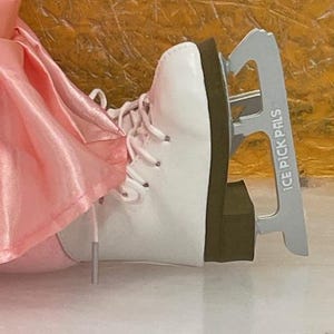 May include: White ice skate with laces and a silver blade. The blade has the text "ICE PICK PALS" printed on it. The skate is next to a pink fabric.