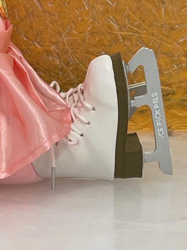 May include: White ice skate with laces and a silver blade. The blade has the text "ICE PICK PALS" printed on it. The skate is next to a pink fabric.