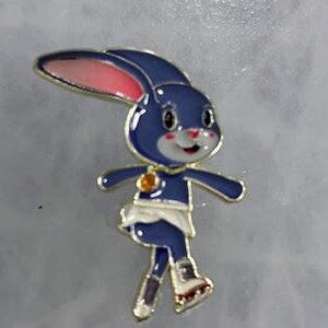 May include: A blue bunny pin with a gold outline, pink inner ears, and a white face. The bunny is wearing a white skirt and gold boots. A gold charm is on the bunny's chest. The pin is on a gray background.
