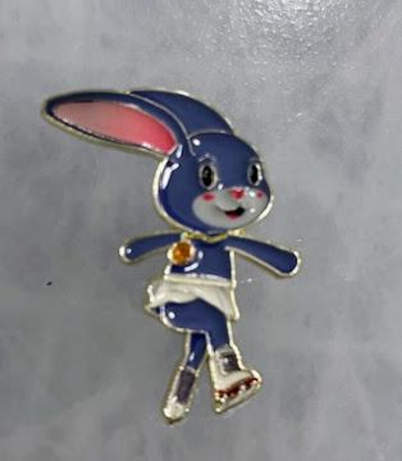 May include: A blue bunny pin with a gold outline, pink inner ears, and a white face. The bunny is wearing a white skirt and gold boots. A gold charm is on the bunny's chest. The pin is on a gray background.