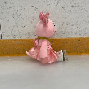 May include: A pink plush axolotl toy wearing a pink skirt and ice skates, sitting on an ice rink. The axolotl has pink frills and a yellow scarf. The background is a white wall with a yellow border.