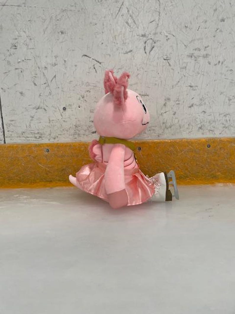 May include: A pink plush axolotl toy wearing a pink skirt and ice skates, sitting on an ice rink. The axolotl has pink frills and a yellow scarf. The background is a white wall with a yellow border.
