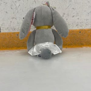 May include: A gray plush bunny toy with long ears, a gold collar, and a white tutu. The bunny has a small, fluffy tail and a colorful bow on its head. The toy is sitting on a light-colored surface.