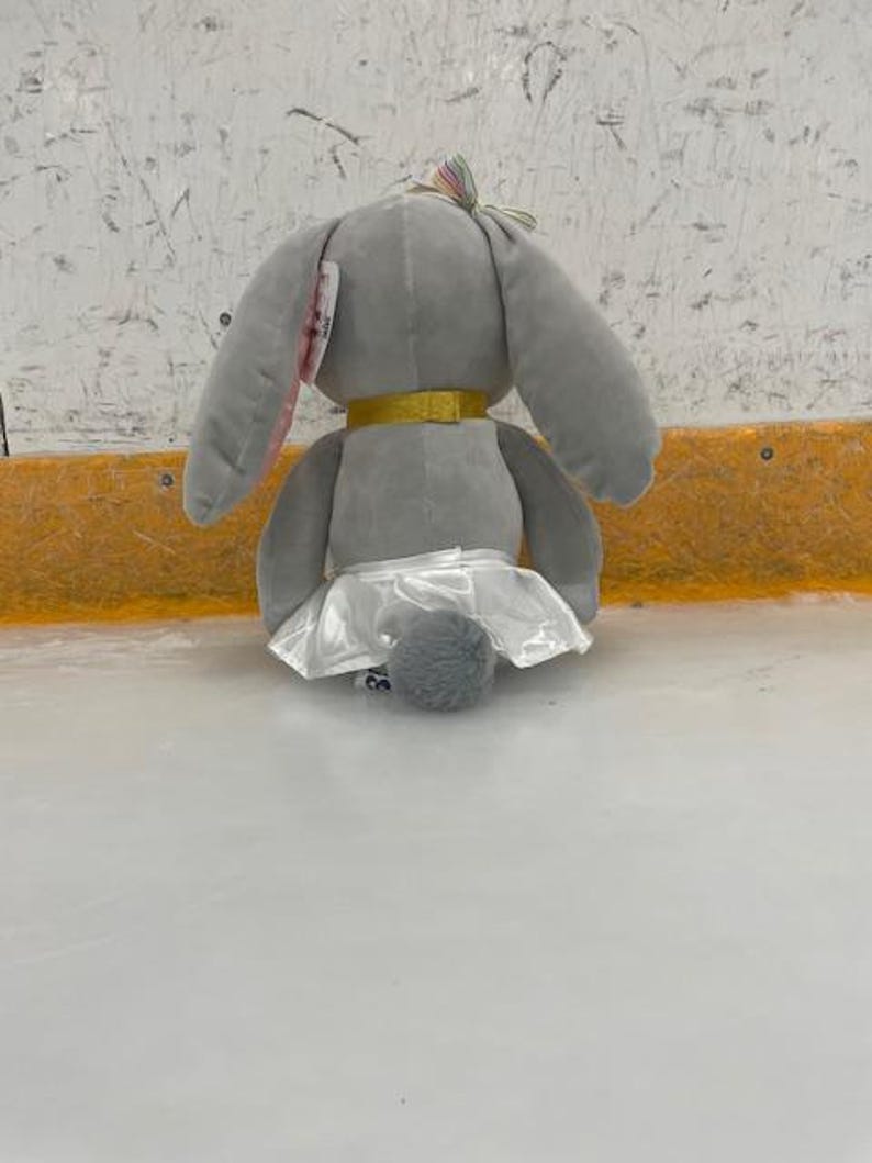 May include: A gray plush bunny toy with long ears, a gold collar, and a white tutu. The bunny has a small, fluffy tail and a colorful bow on its head. The toy is sitting on a light-colored surface.