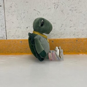 May include: A plush turtle toy wearing a yellow collar and a striped shirt, sitting on an ice rink. The turtle has ice skates on its feet and is wearing pink and white socks. The background is a white wall with a yellow border.