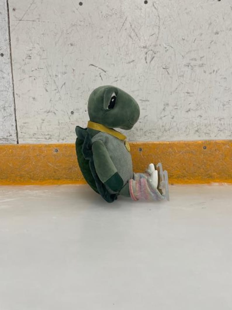 May include: A plush turtle toy wearing a yellow collar and a striped shirt, sitting on an ice rink. The turtle has ice skates on its feet and is wearing pink and white socks. The background is a white wall with a yellow border.