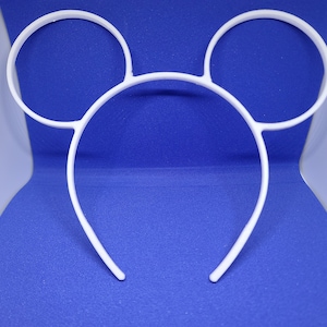 May include: A white headband with two large circular ears on top, resembling a cartoon character. The headband is made of a solid, smooth material and is set against a blue background.