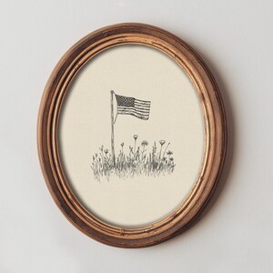 Könnte beinhalten: Ein ovaler Holzrahmen mit einer schwarz-weißen Strichzeichnung auf cremefarbenem Hintergrund. Die Illustration zeigt eine einzelne amerikanische Flagge an einem Mast, die aus einem Feld mit Wildgras und kleinen Wiesenblumen herausragt.