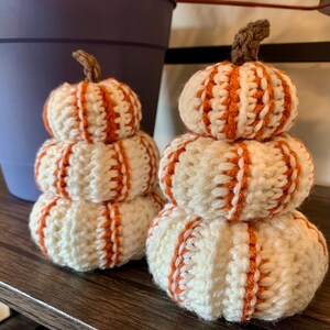 May include: Two crocheted pumpkin stacks, each with three pumpkins. The pumpkins are white with orange stripes. The pumpkins are on a wooden shelf.