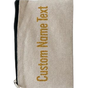 May include: A rectangular beige canvas pouch featuring a black zipper closure on the left side. The front of the pouch displays the text Custom Name Text printed vertically in a glittery gold font, centered on the textured fabric surface.