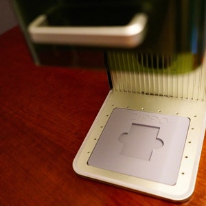 May include: A close-up of a silver and black Zippo lighter with a gray base and a cutout for a lighter insert. The lighter is on a wooden surface.