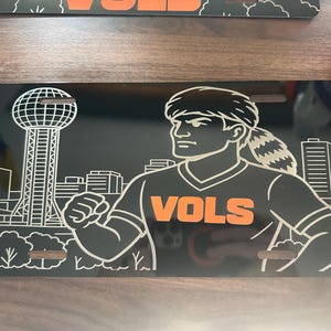 Puede incluir: Placa de matrícula negra con una ilustración estilizada de una persona con una camiseta con la palabra "VOLS" en naranja. El fondo incluye un paisaje urbano y la Sunsphere. La placa tiene tres ranuras para el montaje.