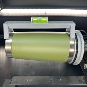Könnte beinhalten: Ein grauer 3D-gedruckter Halter mit einer Wasserwaage oben, der einen grünen Becher in einer Maschine hält.