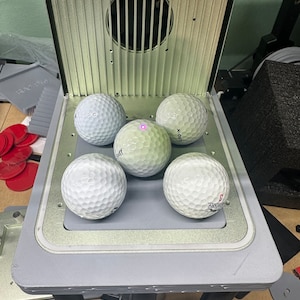 Pode incluir: Cinco bolas de golfe brancas em uma plataforma cinza com uma moldura prateada. A plataforma está em um suporte preto com três pernas. As bolas de golfe estão dispostas em círculo com uma no centro.