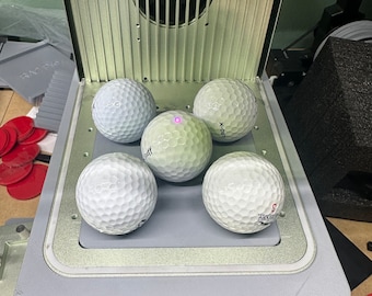 May include: Five white golf balls on a gray platform with a silver frame. The platform is on a black stand with three legs. The golf balls are arranged in a circle with one in the center.