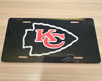 May include: Black license plate featuring the Kansas City Chiefs logo. The logo is a white arrowhead shape with the letters "KC" in red. The license plate has four mounting holes.