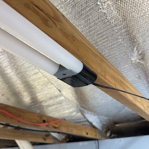 May include: A close-up of a white fluorescent light fixture mounted to a wooden beam. The fixture has a black wire attached to it.