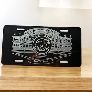 Op de afbeelding: Zwarte nummerplaat met een witte omtrek van een honkbalstadion en een berenlogo. De plaat heeft vier sleuven voor montage en wordt weergegeven op een houten oppervlak. Een sportartikel.