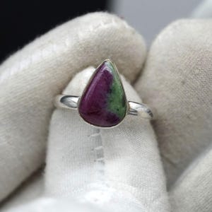 Könnte beinhalten: Ein Silberring mit einem tropfenförmigen Edelstein. Der Edelstein zeigt eine Mischung aus tiefroten und grünen Farbtönen. Der Ring hat ein schlichtes, poliertes Silberband.