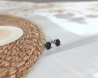 May include: A pair of small, hexagonal stud earrings. The earrings have a dark, textured appearance and are set in silver-colored metal. They are displayed on a white surface with a woven, tan-colored placemat in the background.