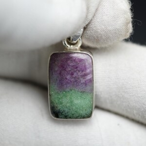 Könnte beinhalten: Ein rechteckiger Anhänger mit silberfarbener Fassung. Der Stein ist in zwei Bereiche unterteilt: ein oberer Bereich in Lila und ein unterer Bereich in Grün. Der Anhänger wird von einer behandschuhten Hand gehalten.