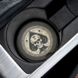 May include: A black and white car coaster with a skull and ace of spades design. The coaster is in a car cup holder.