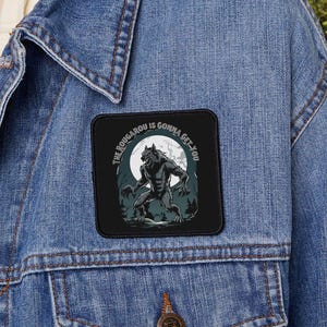 May include: A denim jacket with a black patch featuring a werewolf illustration under a full moon. The patch has the text "The Rougarou is Gonna Get You" in a circular font. The jacket is blue.