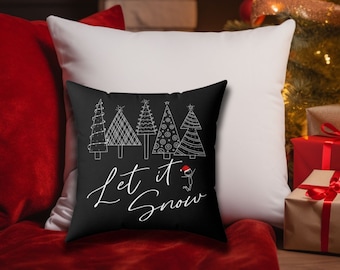 May include: A black decorative pillow with white line art Christmas trees and the phrase "Let it Snow" in white script. The pillow is placed on a red surface with a white pillow behind it. A Christmas tree and gifts are in the background.