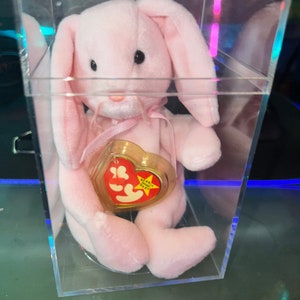 Può includere: Peluche di coniglio rosa Beanie Baby in una teca di plastica trasparente. Il coniglio ha un nastro rosa intorno al collo e un'etichetta a forma di cuore che recita "Beanie Babies Baby".