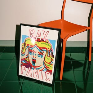 May include: A framed art print with the words "GAY PANIC" in red text. The artwork features a pop art style illustration of a woman with blonde hair, her face showing shock. An orange chair is in the background.