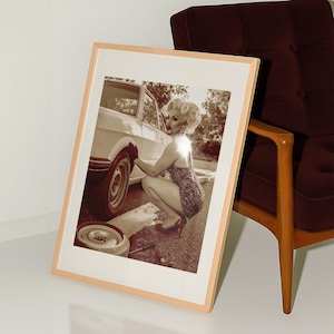May include: Sepia-toned framed print showing a person in a dress and heels changing a car tyre. The image is leaning against a wall next to a maroon armchair with wooden legs. A spare tyre is on the ground.