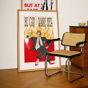 Könnte beinhalten: Ein gerahmter Kunstdruck mit dem Text "BE GAY RAISE HELL" zeigt eine sitzende Figur in Anzug und roten Absätzen vor rotem Hintergrund und Flammen. Ein schwarzer und verchromter Stuhl mit gewebter Sitzfläche befindet sich im Vordergrund.